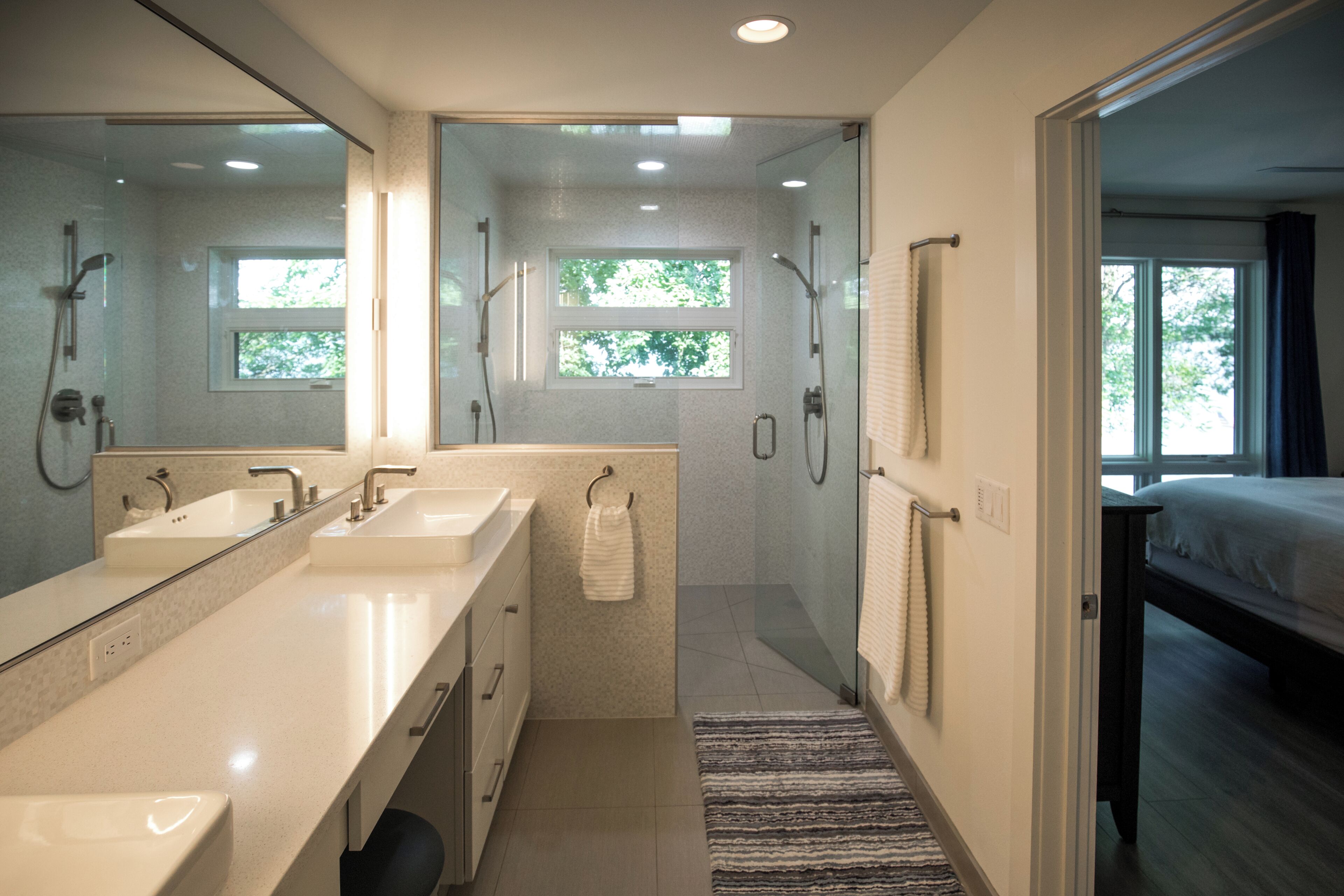 A modern bathroom with two sinks, a glass shower enclosure, and a view into a bedroom with a bed.