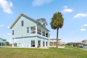 A light blue two-story house with a balcony, a palm tree in the foreground, and a marina in the background.