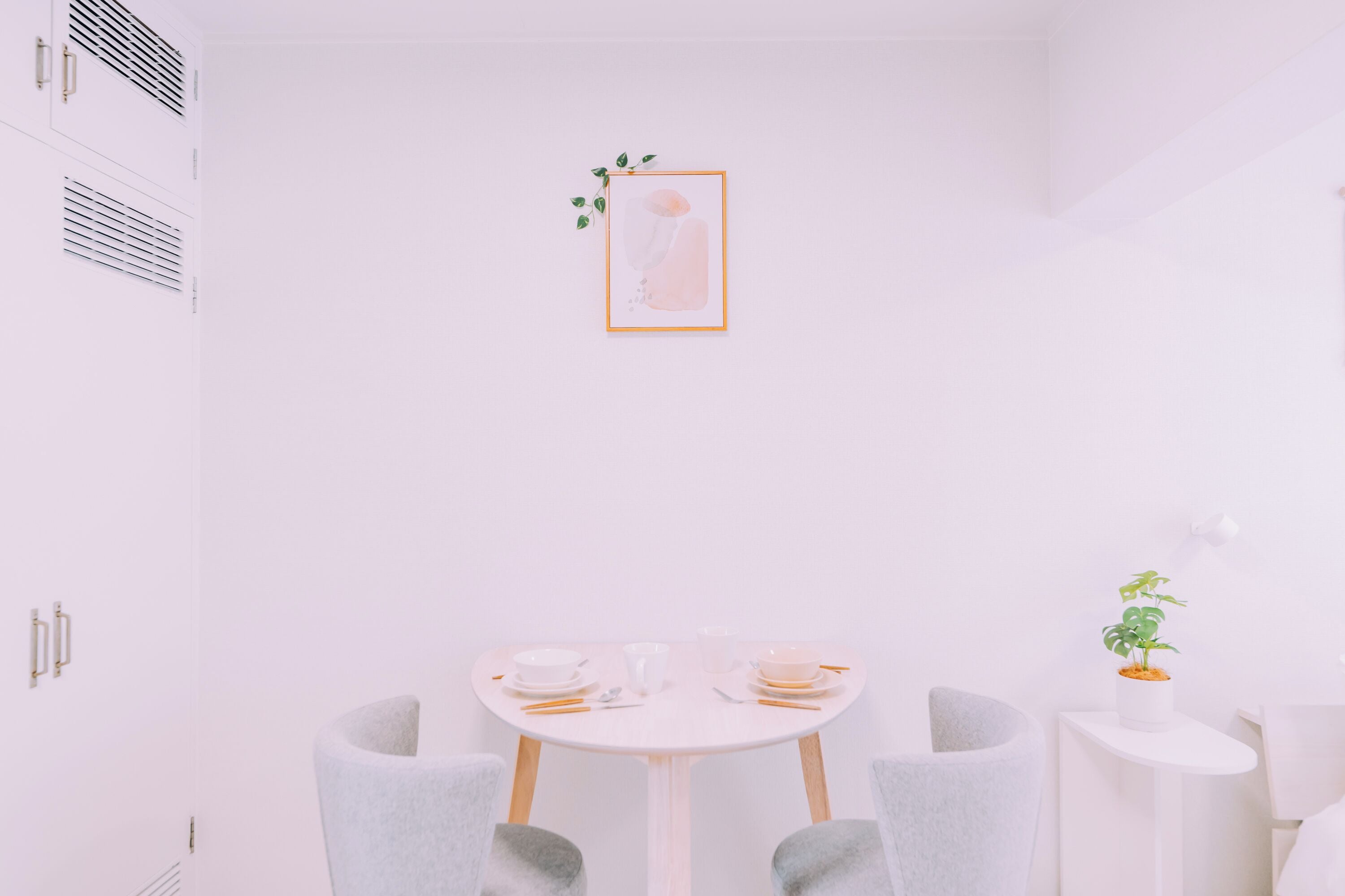 Ein minimalistisch eingerichteter Raum mit einem runden Tisch, zwei Stühlen, einem kleinen Beistelltisch mit Pflanze und einem gerahmten Bild an der Wand.