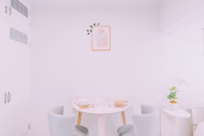 Ein minimalistisch eingerichteter Raum mit einem runden Tisch, zwei Stühlen, einem kleinen Beistelltisch mit Pflanze und einem gerahmten Bild an der Wand.