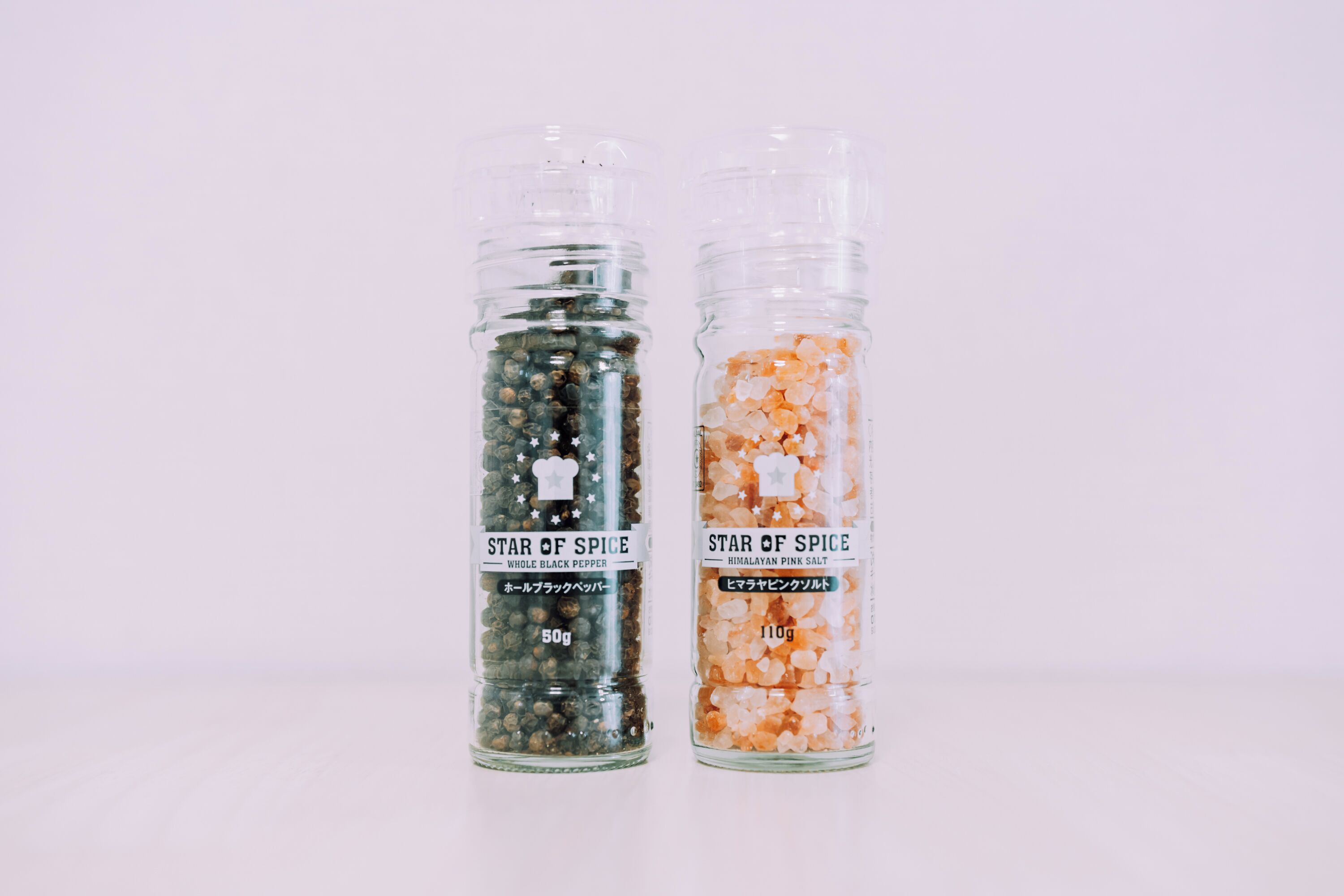 Twee glazen containers met de aanduiding 'Star of Spice', elk gevuld met een ander kruid; één met 'Zwarte Peppercorns' en de andere met 'Roze Himalayazout'.