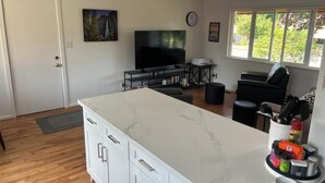 A modern kitchen with a marble countertop, a dining area with a black chair, and a TV in the background. - Awesome Little House ! (Mountlake Terrace)