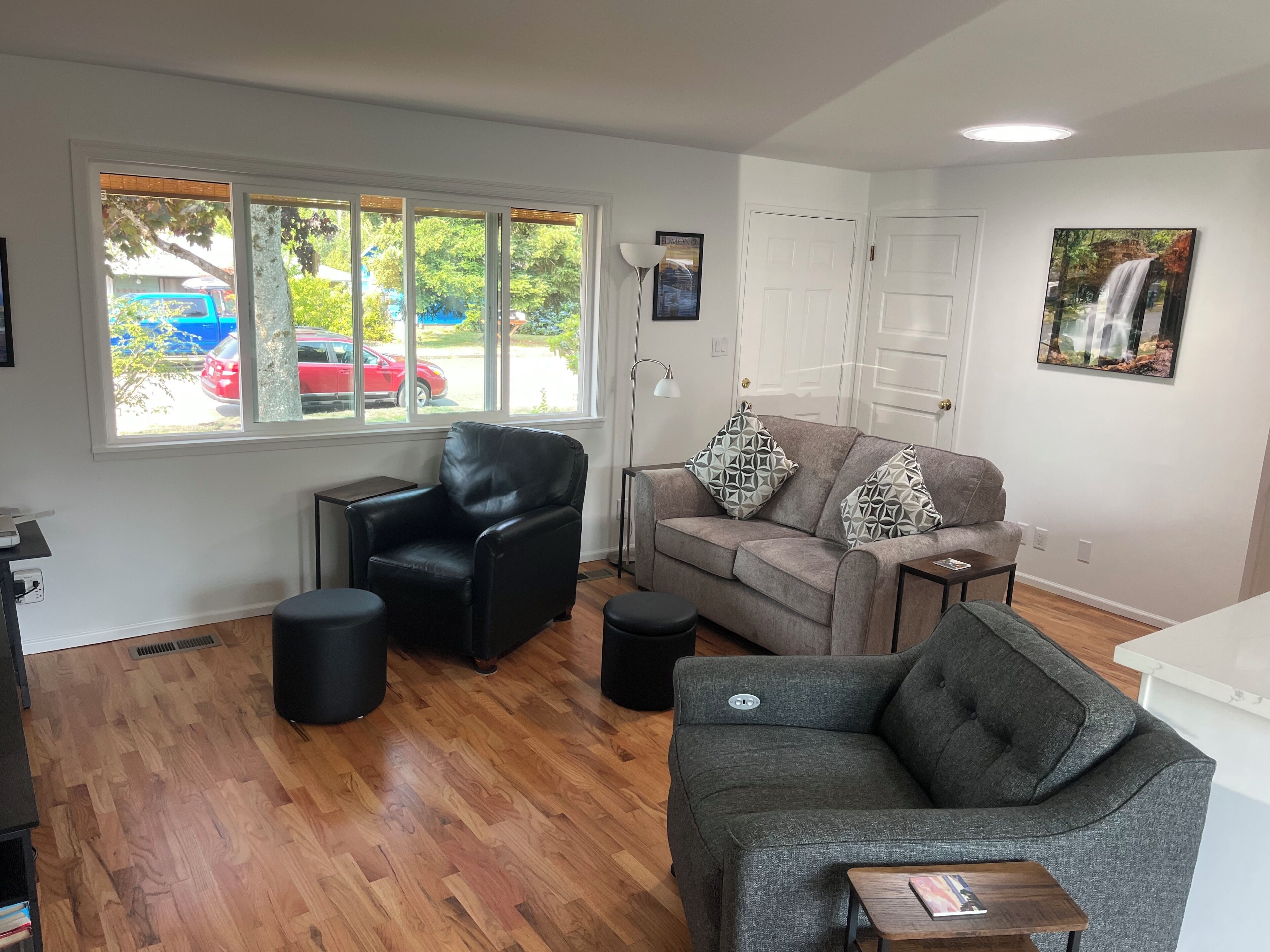 A living room with a leather chair, a grey sofa, and a small table. There is a window with a view of a red car and trees.