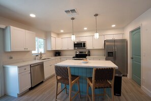 A modern kitchen with white cabinets, stainless steel appliances, and a central island with two chairs. - Unwind in style, home & heated pool for families (Mesa)