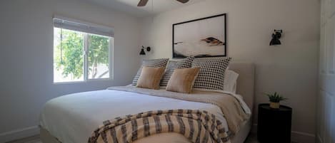 A bedroom with a bed, a ceiling fan, a window, a wall-mounted lamp, and a framed picture.
