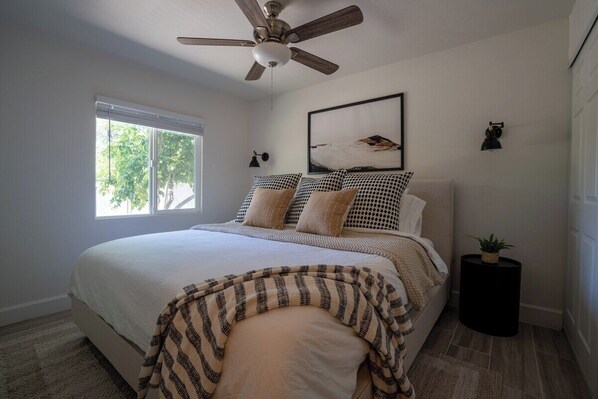 A bedroom with a bed, a ceiling fan, a window, a wall-mounted lamp, and a framed picture.