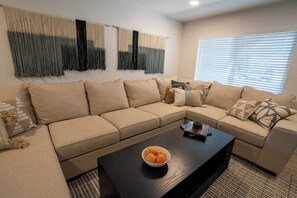 A living room with a beige sectional sofa, a black coffee table with a bowl of fruit, and a window with blinds.
