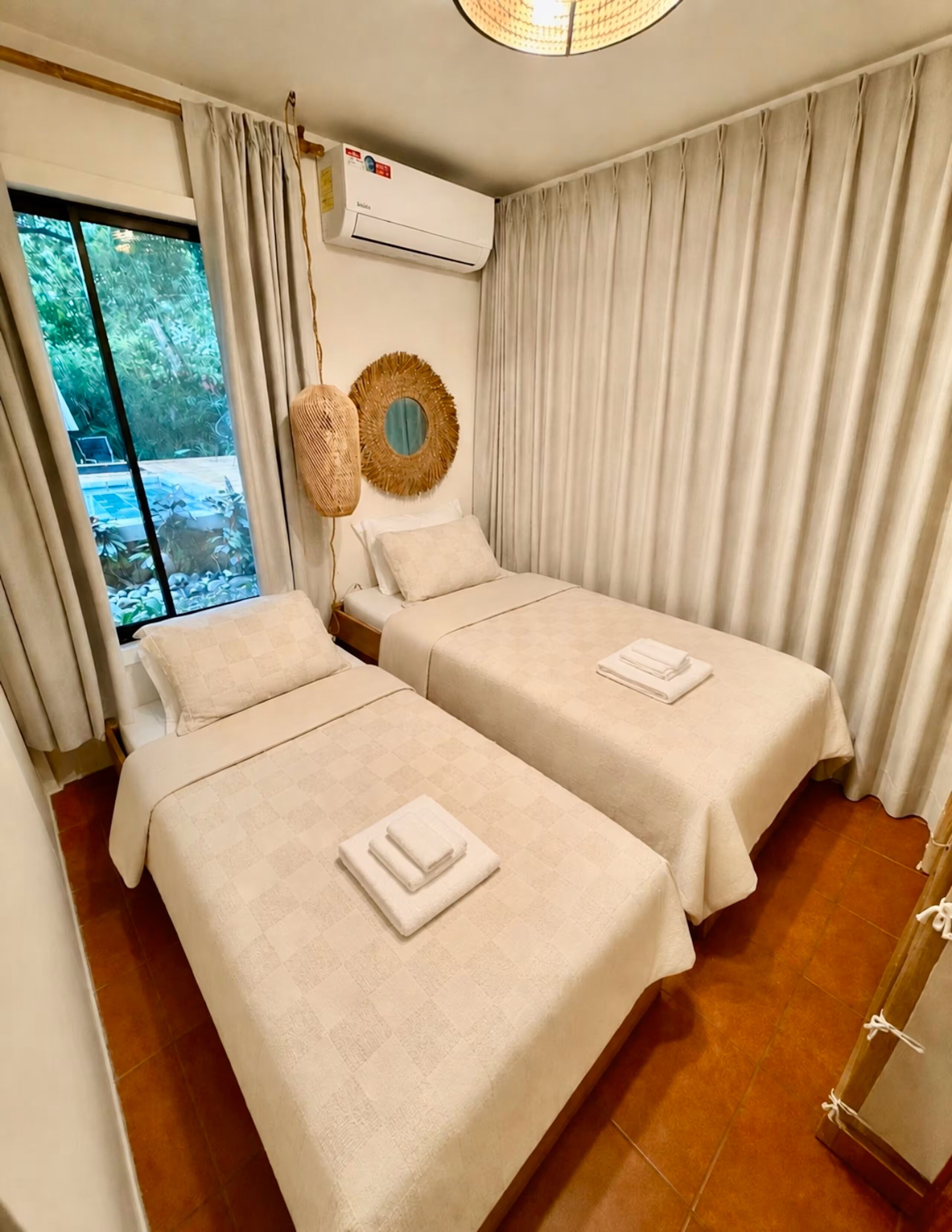 A hotel room with two beds, a large window, an air conditioning unit, and a view of greenery outside.