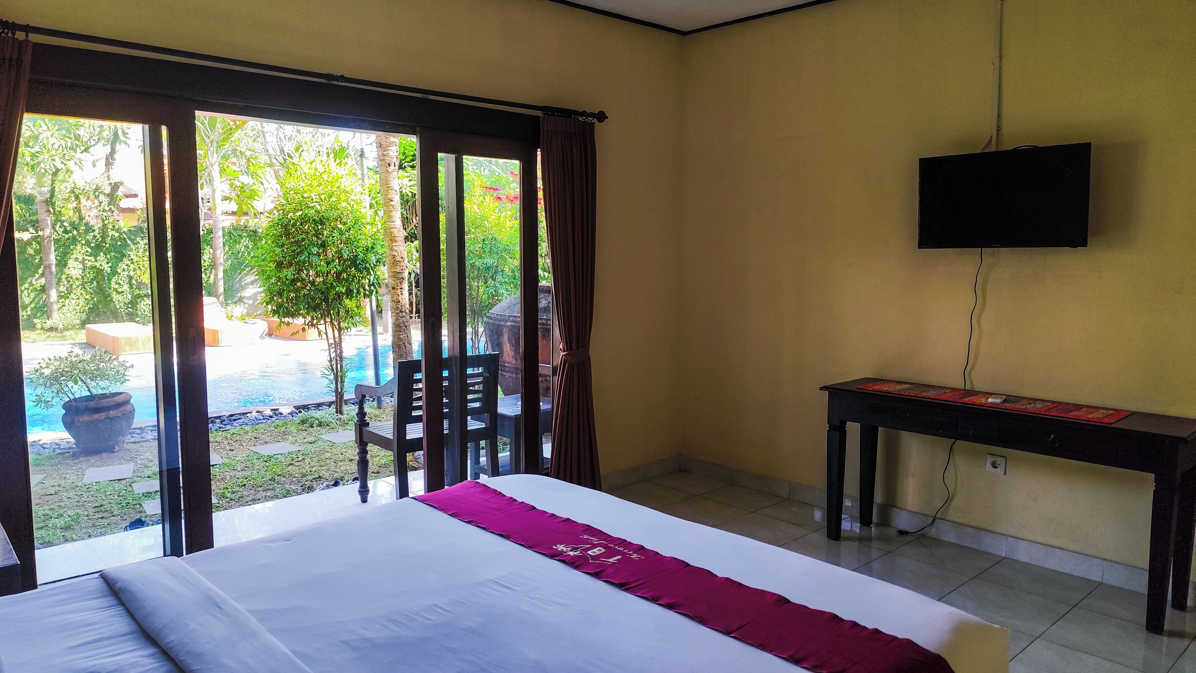 A hotel room with a bed, a desk, a TV, and a view of an outdoor pool area.