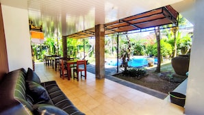 A covered outdoor seating area with tables and chairs, a pool, and lush greenery.