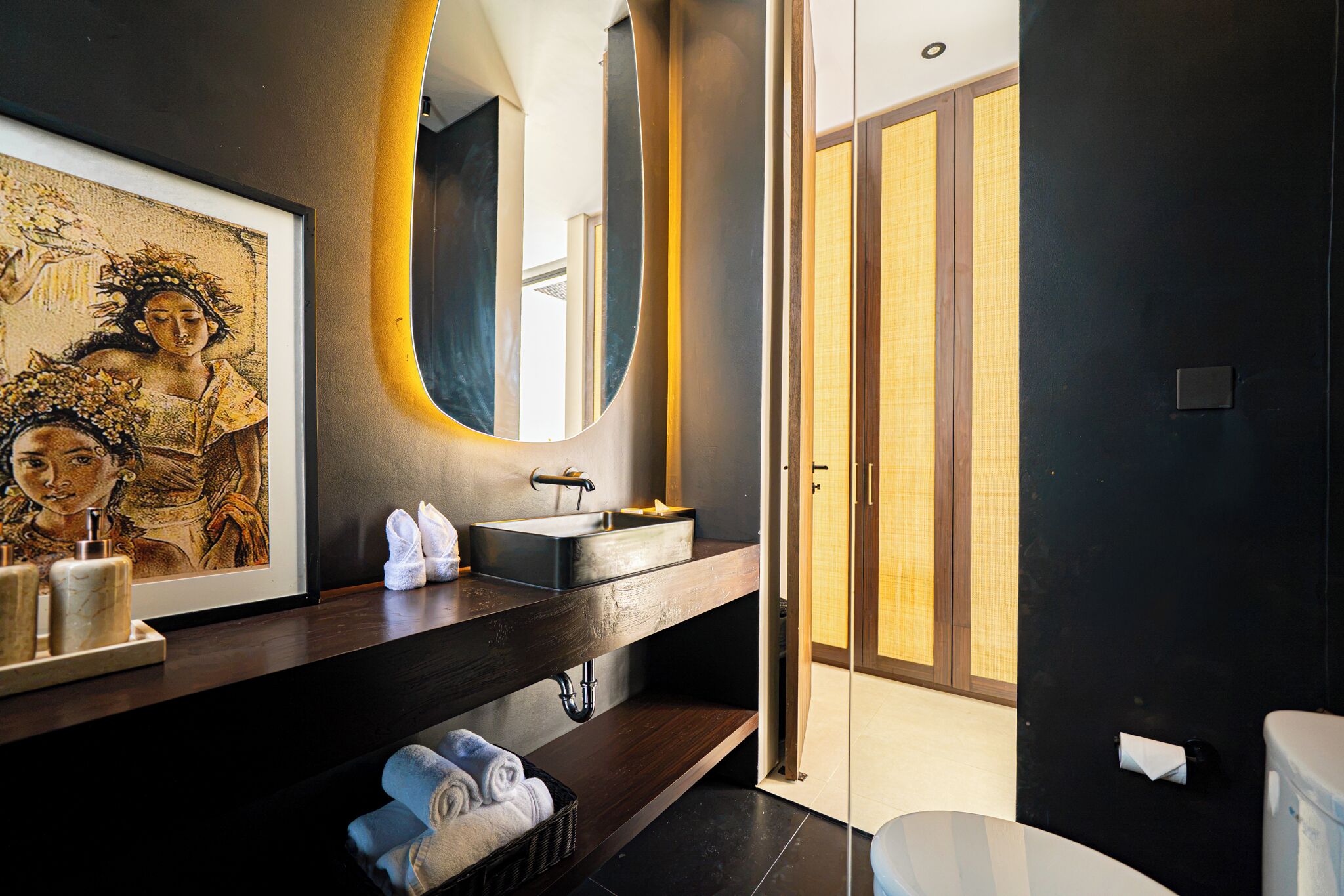 A modern bathroom with a dark wall, a large mirror, a sink, and a toilet. There is a framed picture on the wall and towels on the counter.