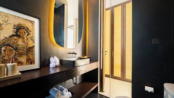 A modern bathroom with a dark wall, a large mirror, a sink, and a toilet. There is a framed picture on the wall and towels on the counter.