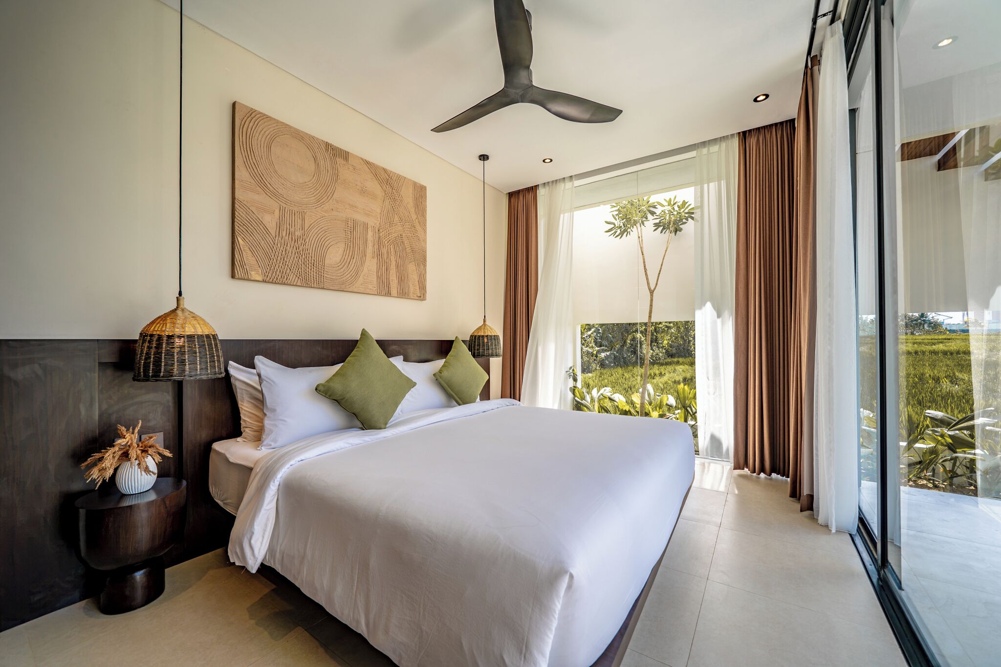 A modern hotel bedroom with a large bed, a ceiling fan, a view of a garden through a sliding door, and a wall-mounted artwork.