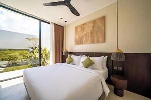A hotel room with a large bed, a view of a green landscape, a ceiling fan, and a wall-mounted artwork. - Villa Ambisi Kedungu (Tabanan)