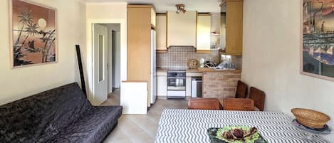 Un espacio habitable compacto con sofá, una mesa de comedor con camino de mesa estampado, zona de cocina con estufa y armarios, y obras de arte en las paredes.