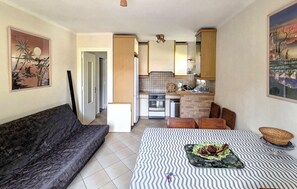 Ein gemütlicher Wohnraum mit Sofa, Esstisch mit einem gemusterten Tischläufer, Küchenzeile mit Herd und Schränken sowie Bildern an den Wänden.