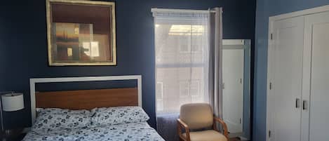 A bedroom with a bed, a chair, a wardrobe, a window with curtains, and a framed picture on the wall.