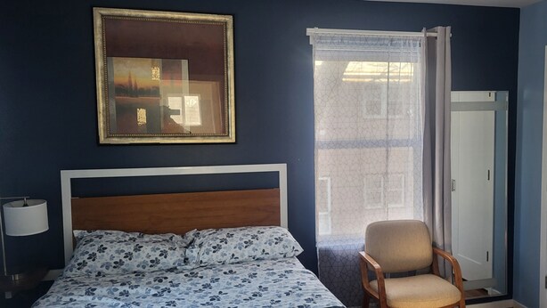 A bedroom with a bed, a chair, a wardrobe, a window with curtains, and a framed picture on the wall.