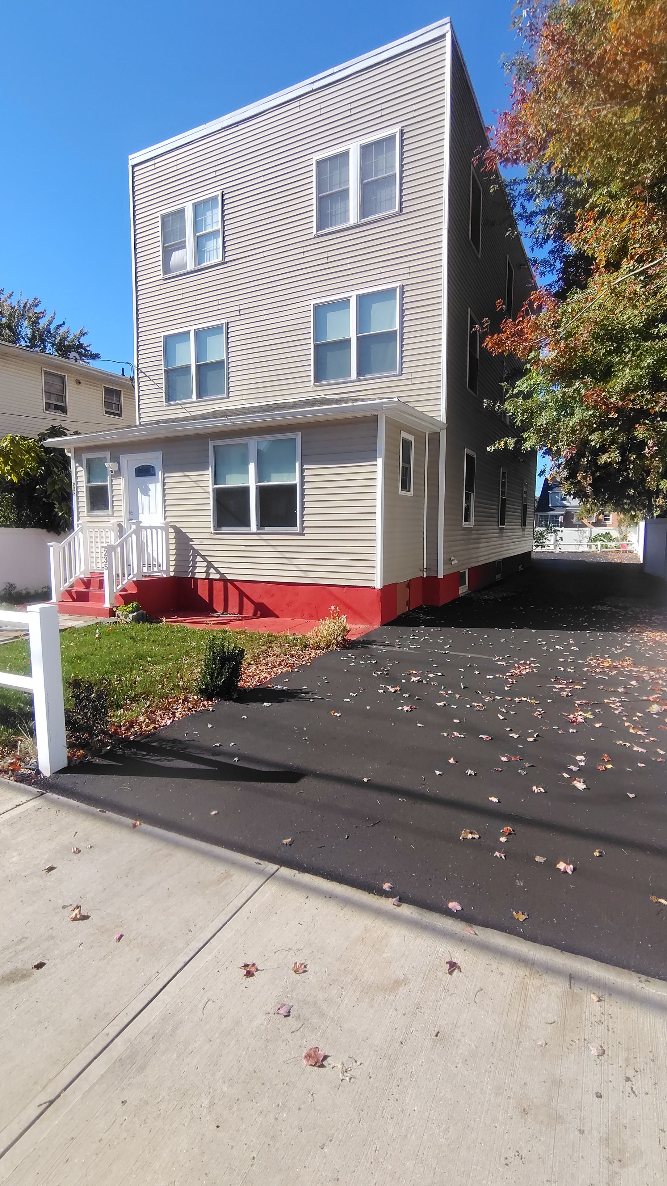 A two-story building with a red base and beige siding, a front porch with a white railing, and a driveway with scattered leaves.