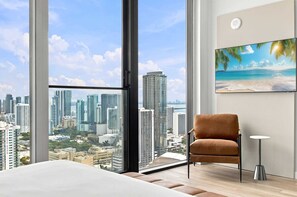 A high-rise room with a large window, a brown chair, a small table, and a painting of a beach scene. - 44th Floor at E11EVEN with Panoramic City Views (Miami)