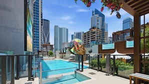Uma piscina no terraço com barreira de vidro, rodeada por edifícios altos e uma escultura colorida.