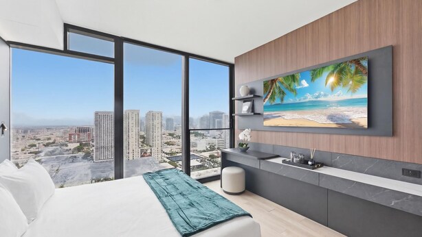 A modern hotel room with a large bed, a view of the city, a kitchen area, and a TV displaying a beach scene. - E11EVEN Studio Suite w/ Rooftop Pool & City Views (Miami)