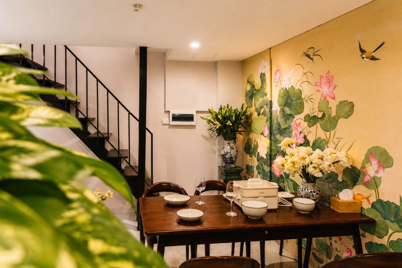 Un comedor con una mesa de madera dispuesta para dos personas, un jarrón con flores y una escalera al fondo.