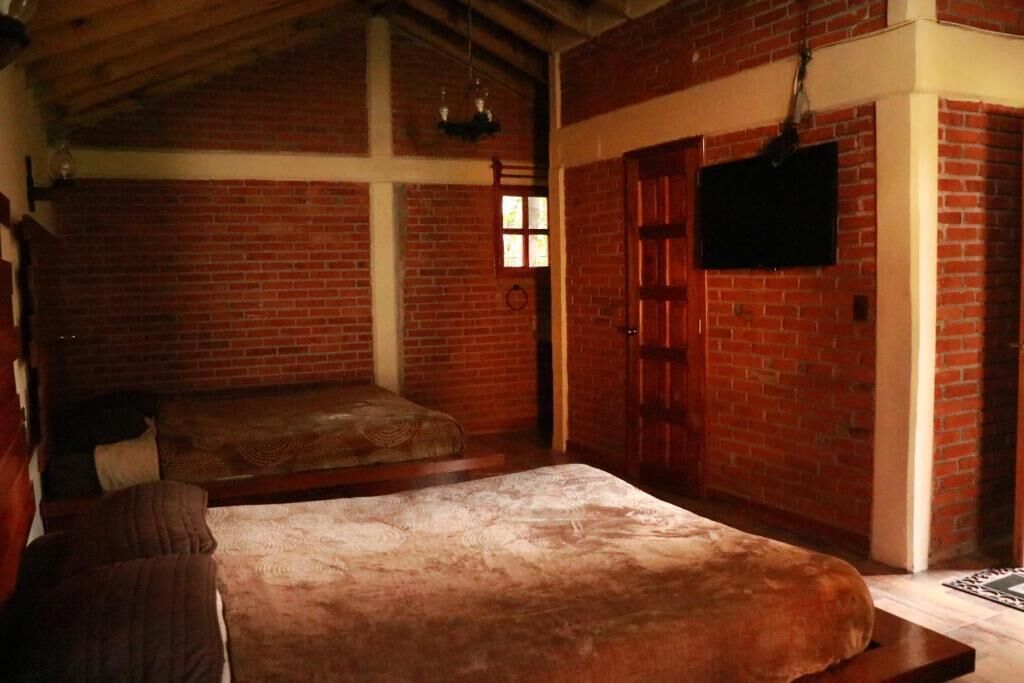 A room with a brick wall, a bed with a patterned bedspread, a wooden bench, and a television mounted on the wall.