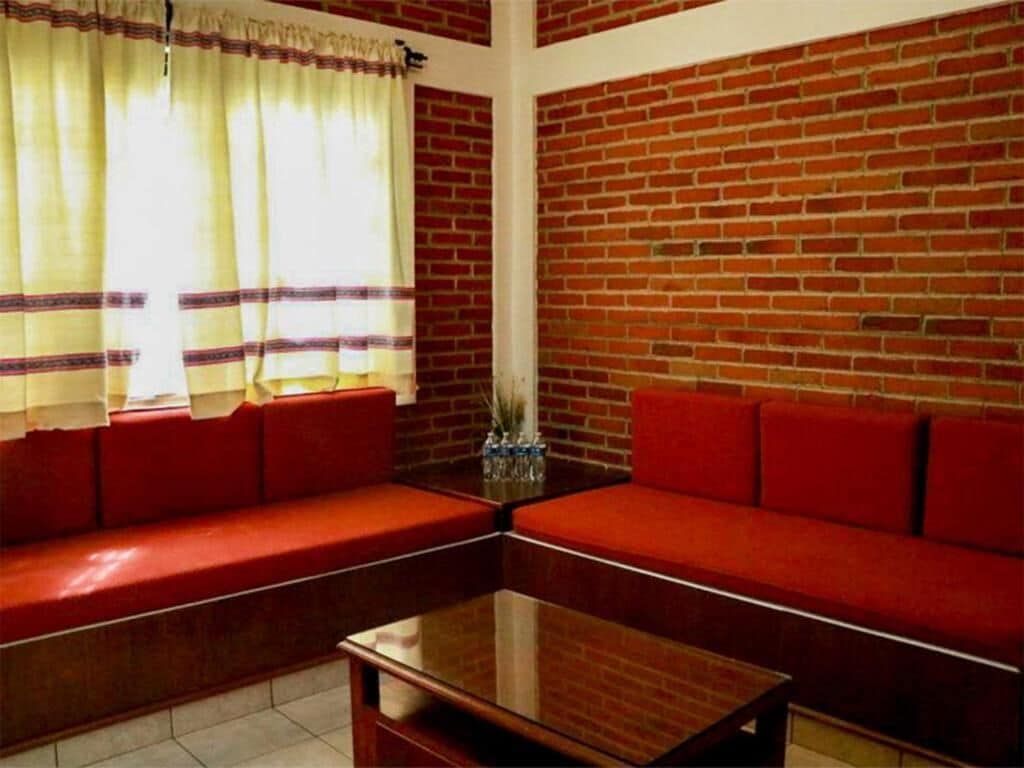 A room with a brick wall, red upholstered seating, a glass coffee table, and a window with curtains.