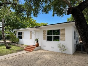 A single-story house with a white exterior, a front porch with steps, a large potted plant, and a tree in the background. - Bright, stylish 3BR/2BA pet-friendly retreat near Wynwood, Design District & MIA (Miami)