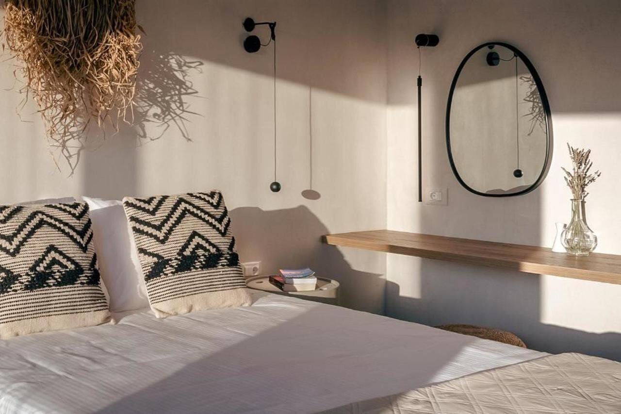 A bedroom with a bed, pillows, a bedside table, a hanging light fixture, a mirror, and a wall-mounted shelf with a vase.