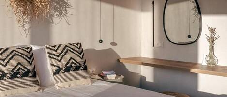 A bedroom with a bed, pillows, a bedside table, a hanging light fixture, a mirror, and a wall-mounted shelf with a vase.