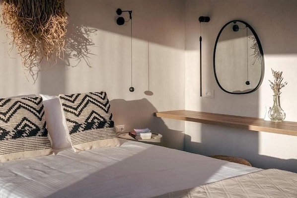 A bedroom with a bed, pillows, a bedside table, a hanging light fixture, a mirror, and a wall-mounted shelf with a vase.