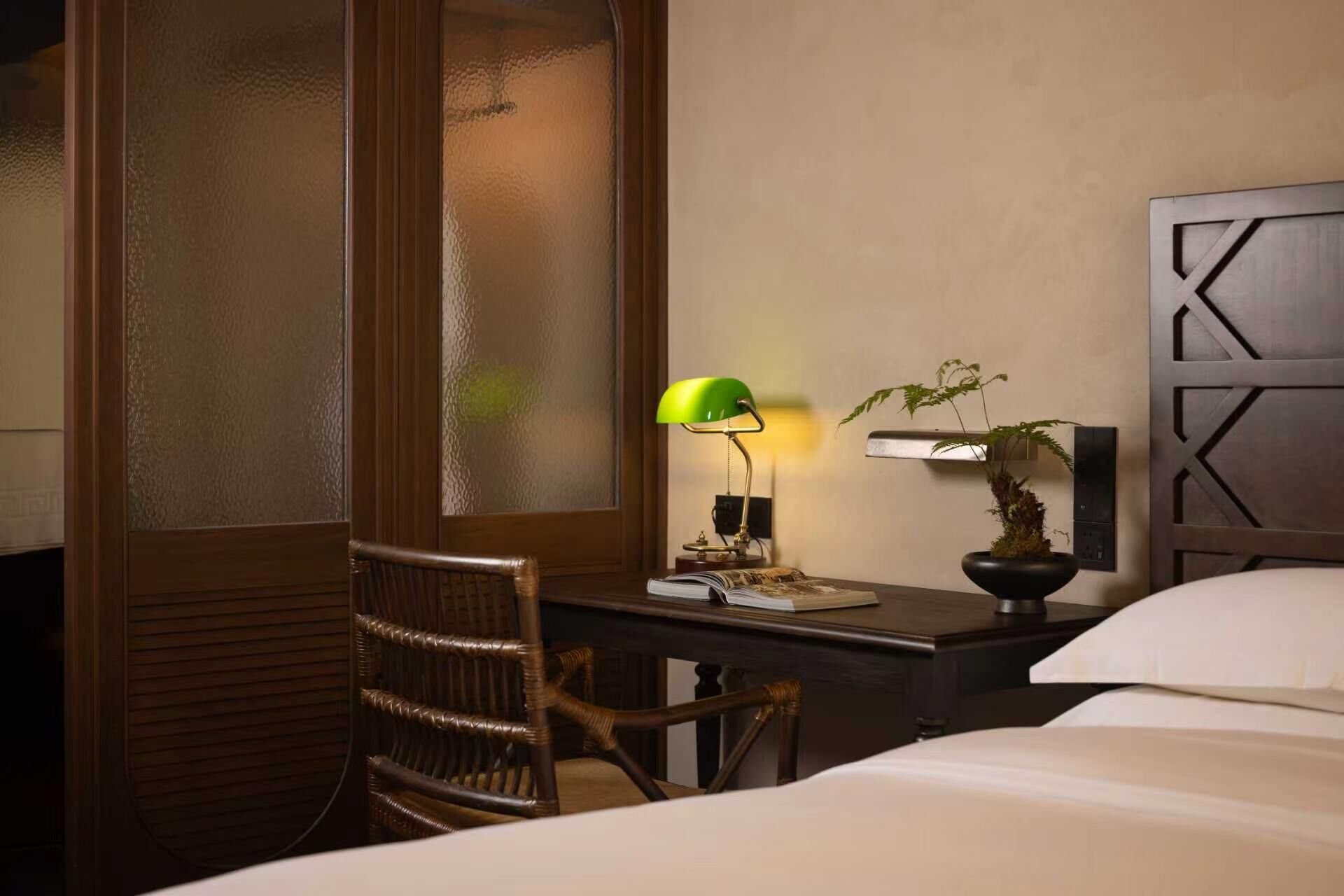 A hotel room with a bed, a desk with a lamp, a chair, a plant, and a wall-mounted light fixture.