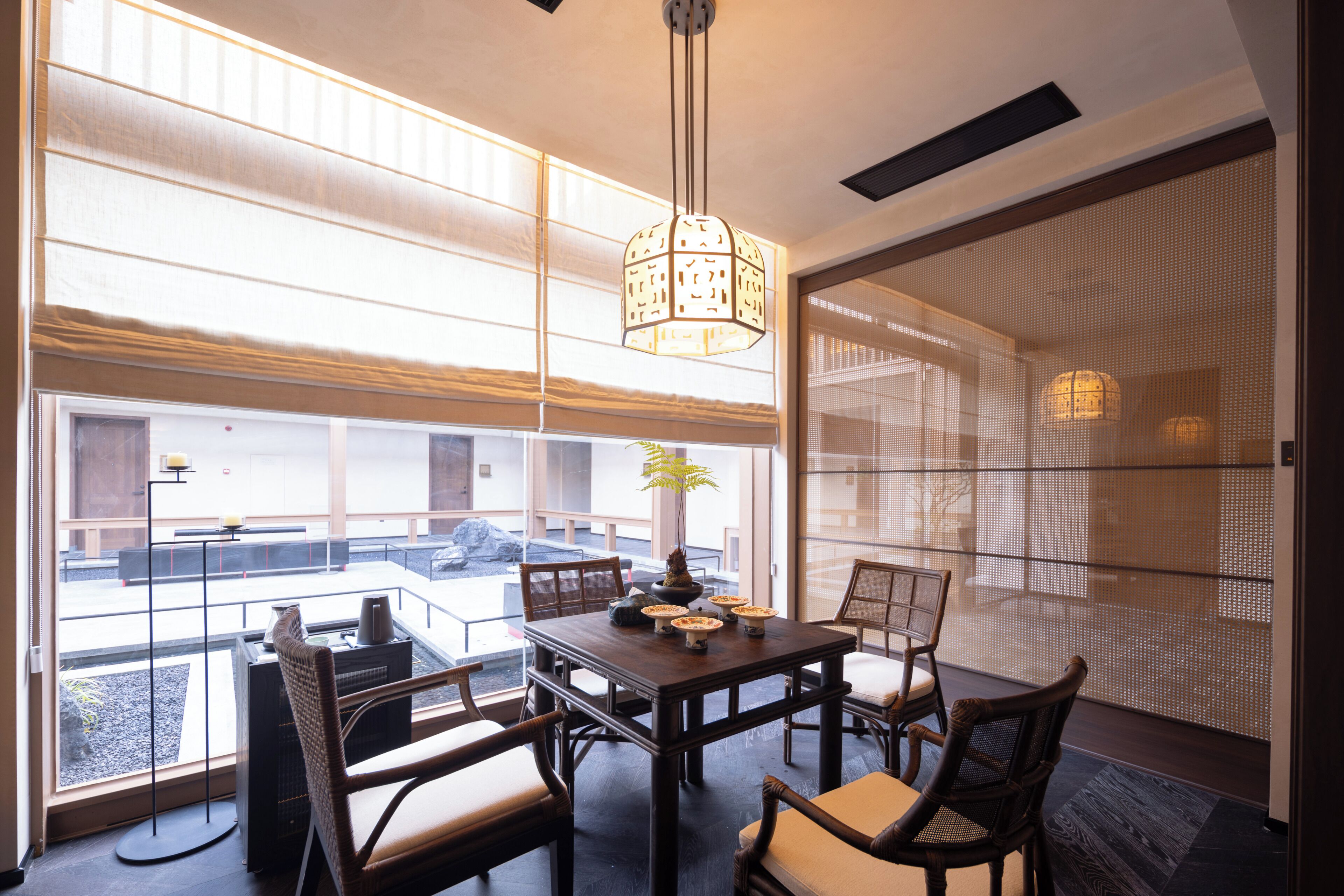 A dining area with a table set for four, chairs, a hanging light fixture, and a view through large windows.