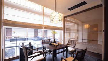 A dining area with a table set for four, chairs, a hanging light fixture, and a view through large windows.