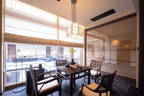 A dining area with a table set for four, chairs, a hanging light fixture, and a view through large windows.