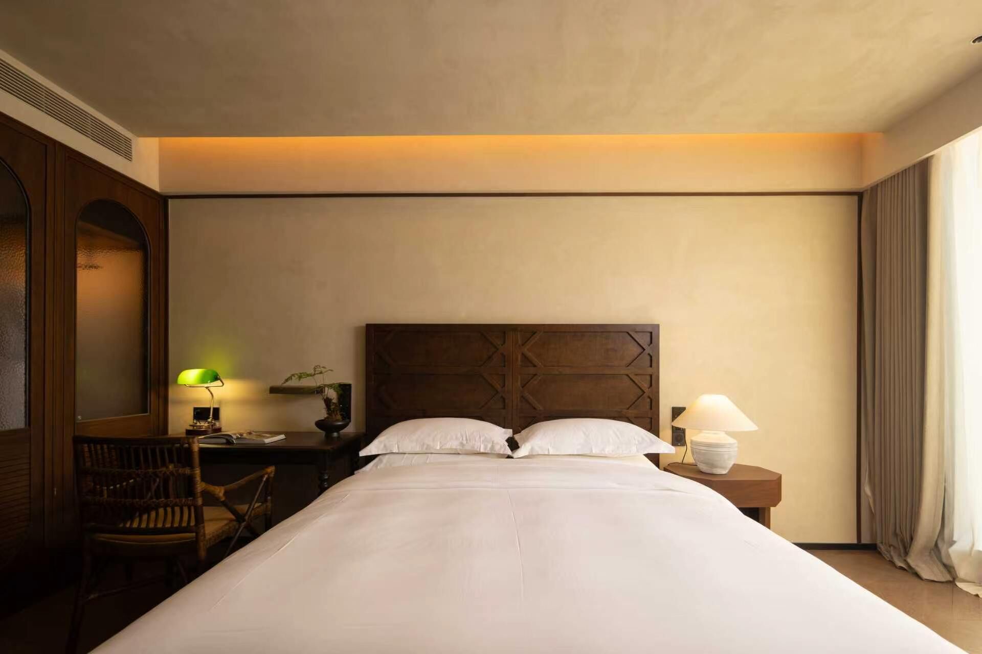 A hotel room with a large bed, a wooden headboard, a nightstand with a lamp, a chair, and a window with curtains.