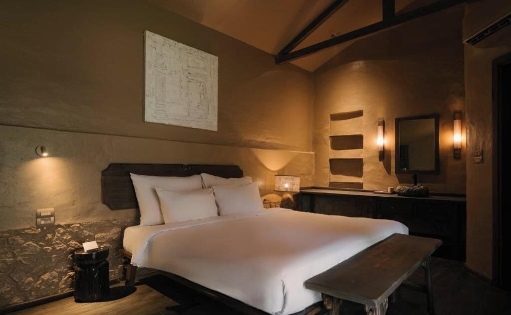 A bedroom with a large bed, bedside tables, a bench, a mirror, and wall-mounted lights.