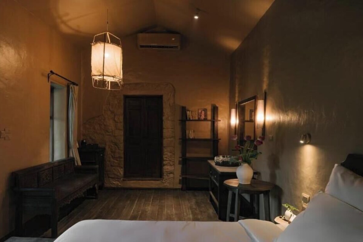A cozy room with a bed, a bench, a small table, a bookshelf, and a stone wall.