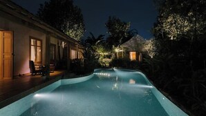 Uma piscina com borda curva, rodeada por um deck de madeira e um edifício com janelas amplas.