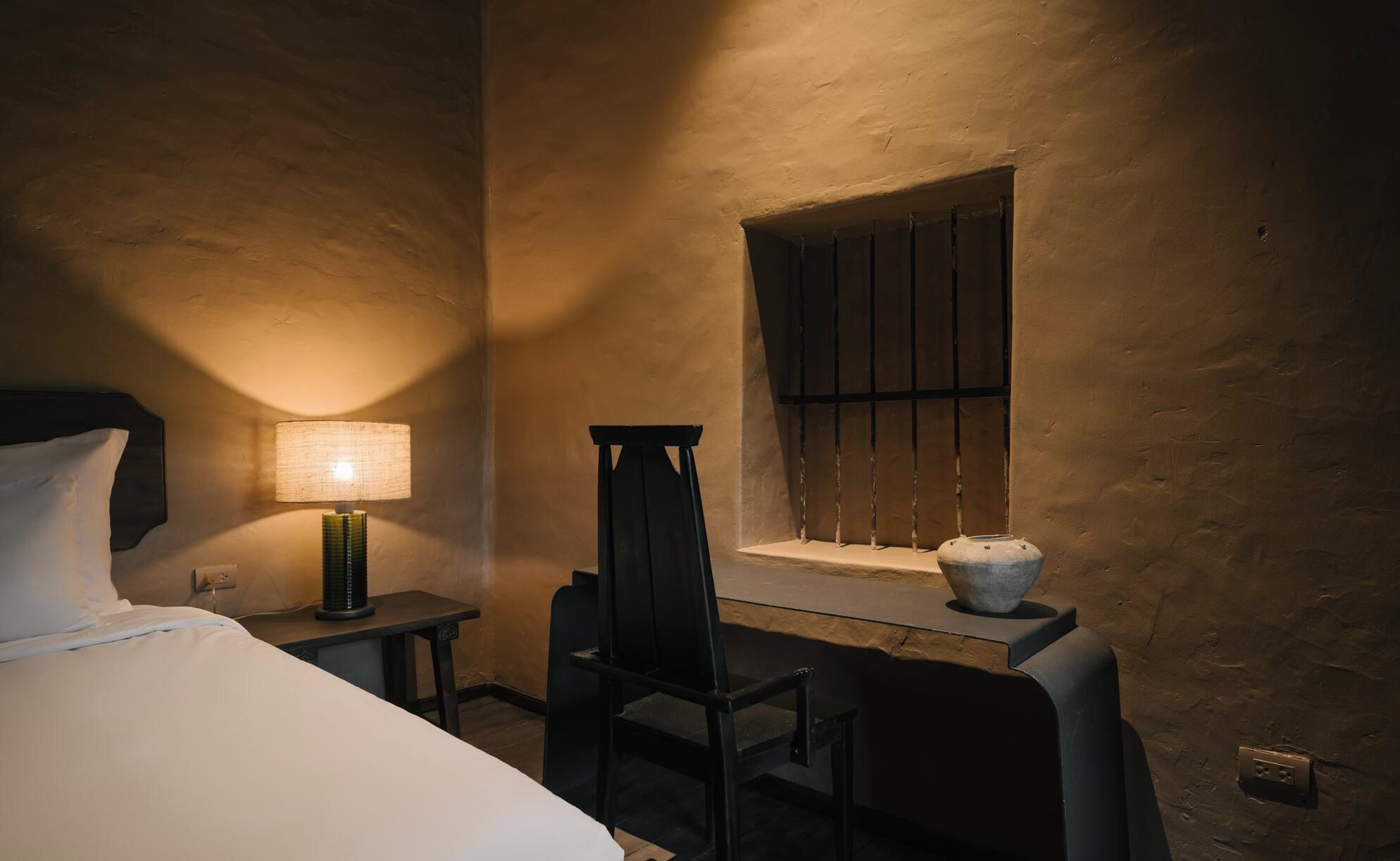 Ein Hotelzimmer mit Bett, Schreibtisch, Stuhl, Lampe und einem Fenster mit Gitter.