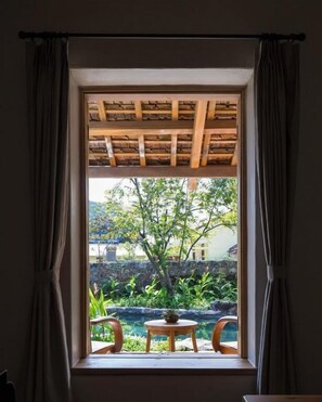 A window with a wooden ceiling structure, a view of a garden with a pool, and a table with chairs outside.