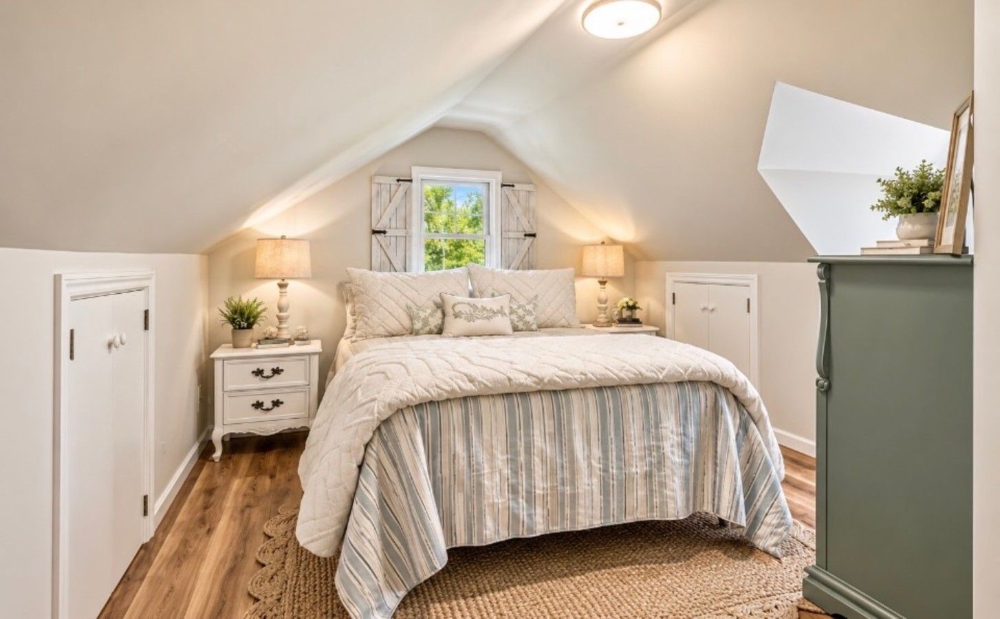 A bedroom with a sloped ceiling, a bed with striped bedding, bedside lamps, and a view of greenery outside.