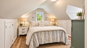 A bedroom with a sloped ceiling, a bed with striped bedding, bedside lamps, and a view of greenery outside.