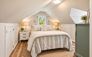 A bedroom with a sloped ceiling, a bed with striped bedding, bedside lamps, and a view of greenery outside.