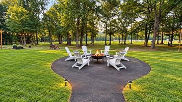 A circular outdoor seating area with a fire pit surrounded by white chairs, set in a grassy field with trees in the background.