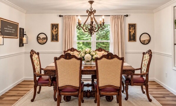A dining room with a wooden table, four chairs, a chandelier, and a window with curtains.