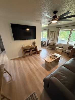Ein Wohnzimmer mit Flachbildfernseher, Holzmöbeln, Deckenventilator und großen Fenstern.