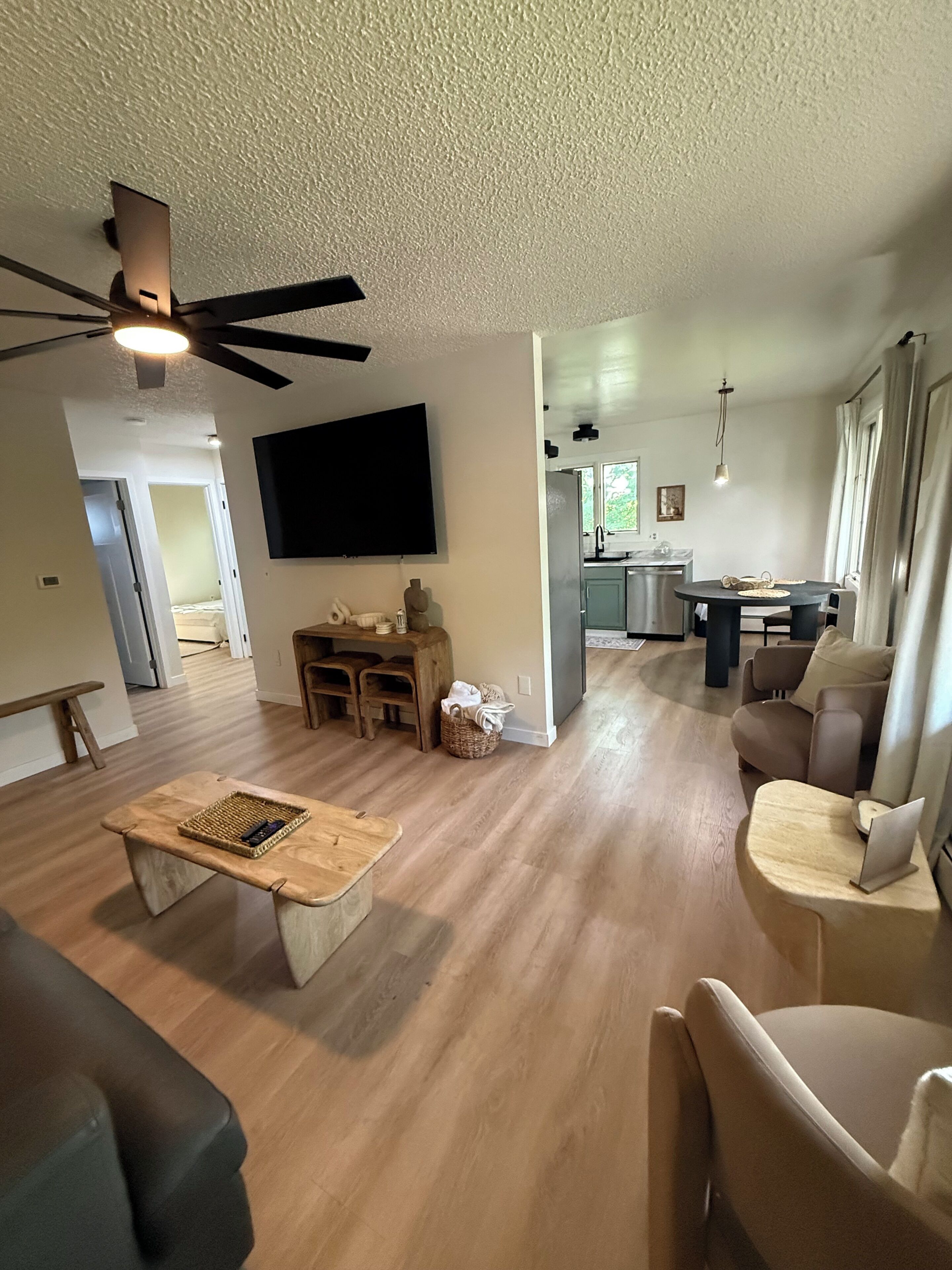 A spacious living room with a ceiling fan, a flat-screen TV mounted on the wall, and a dining area with a round table.